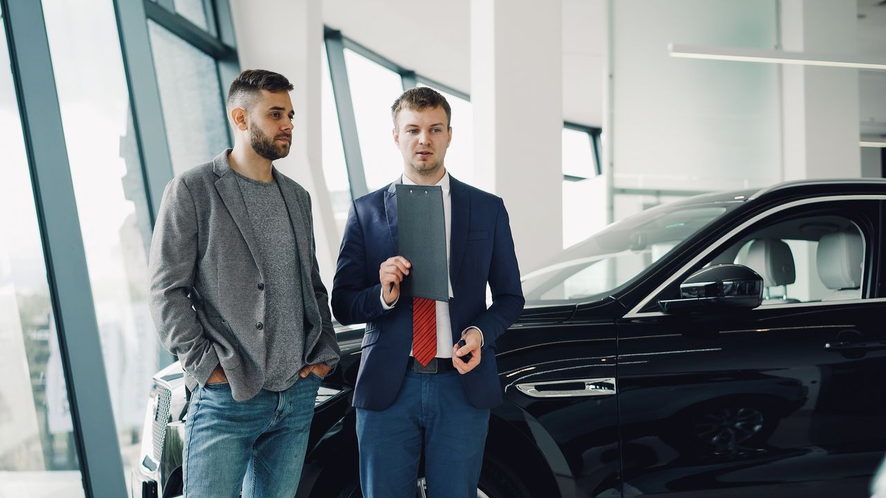 Two businessmen discussing car purchase in modern dealership showroom.